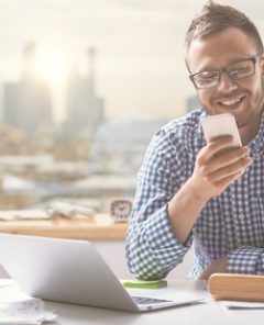 Portrait of handsome european man using cellphone while sitting at office table with laptop computer and other items